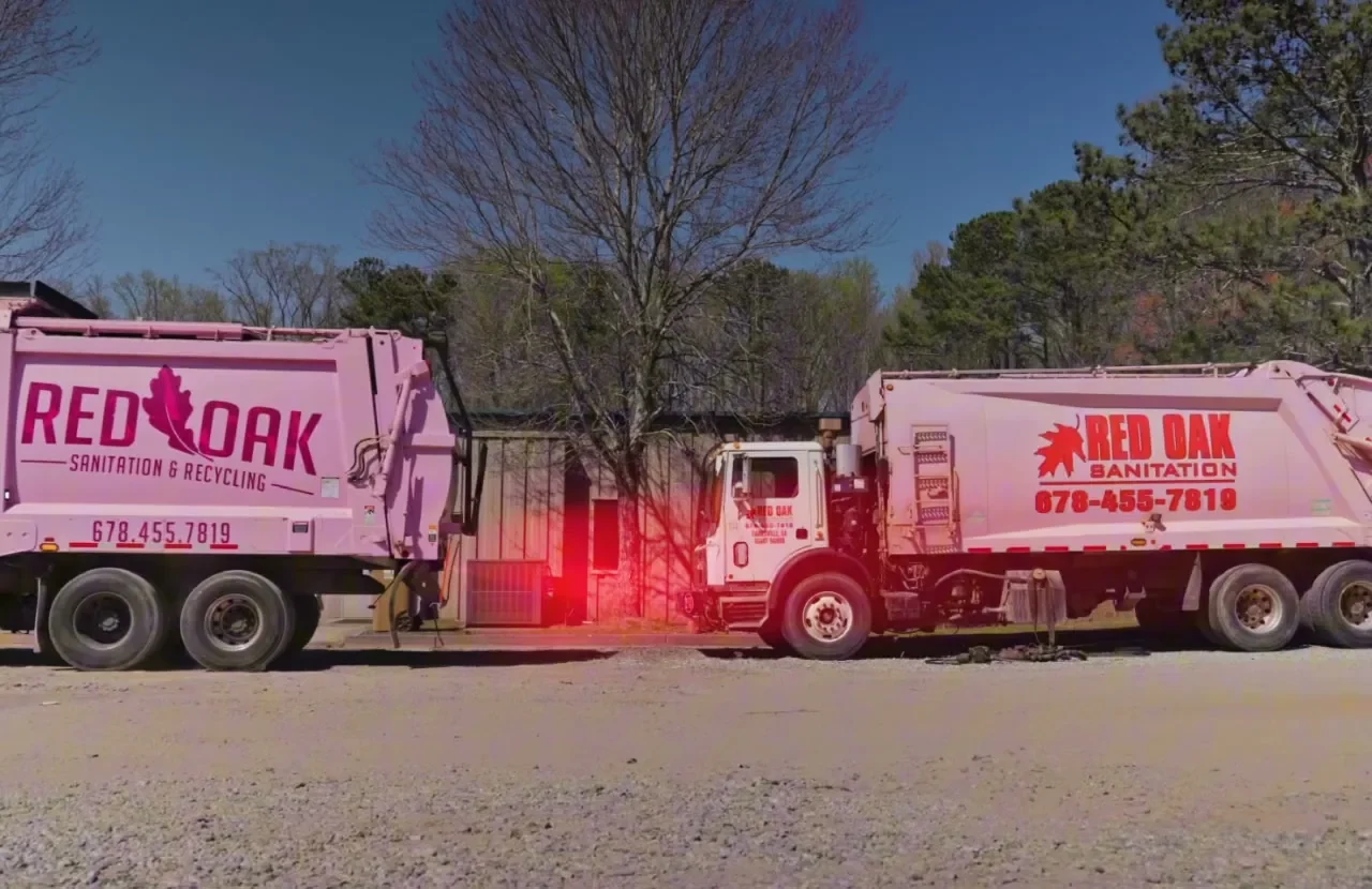 Two Red Oak Sanitation garbage trucks parked in a yard, one with its brake light on