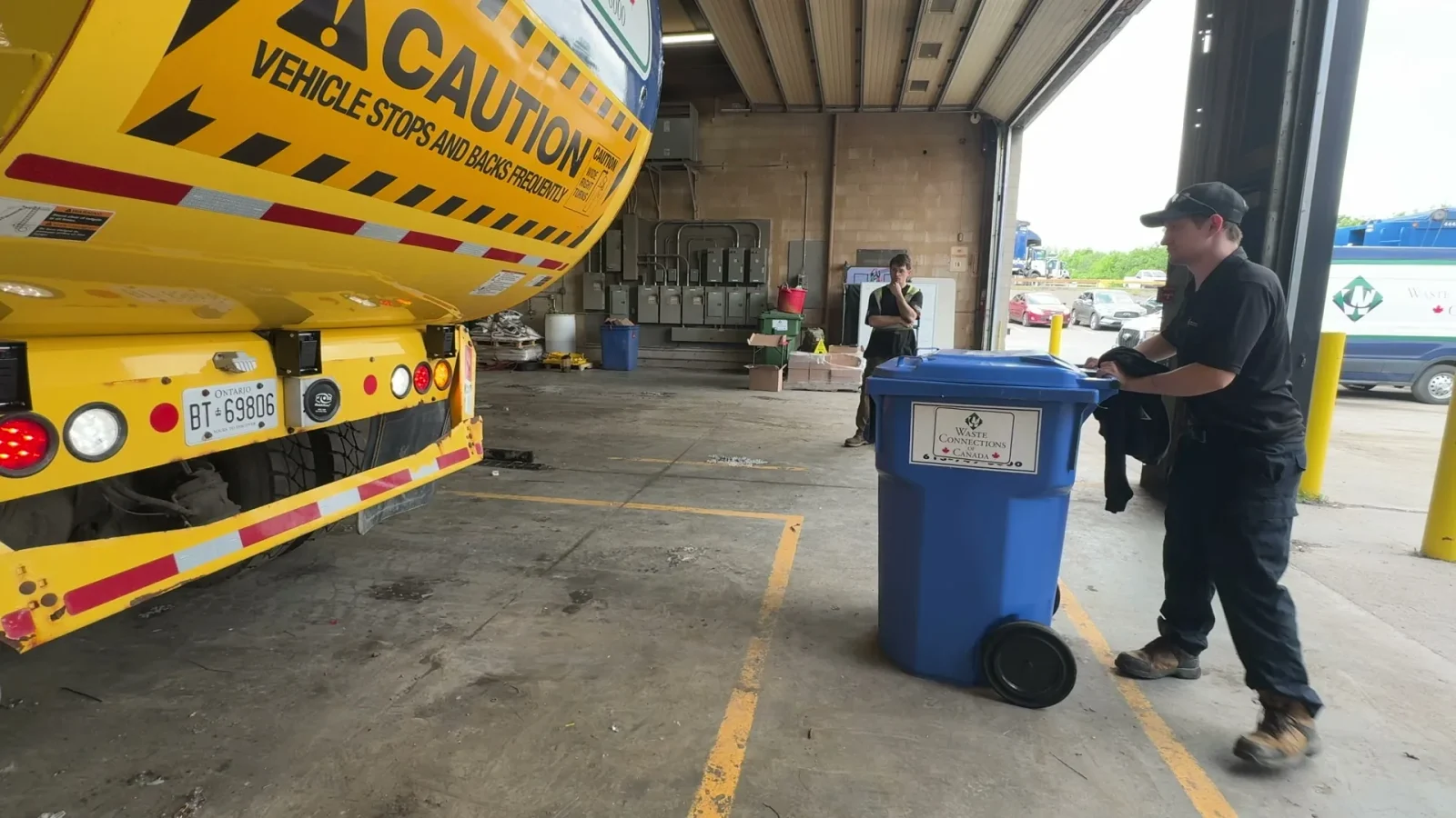 Technician moving a recycling cart behind a yellow front-load truck in a service bay