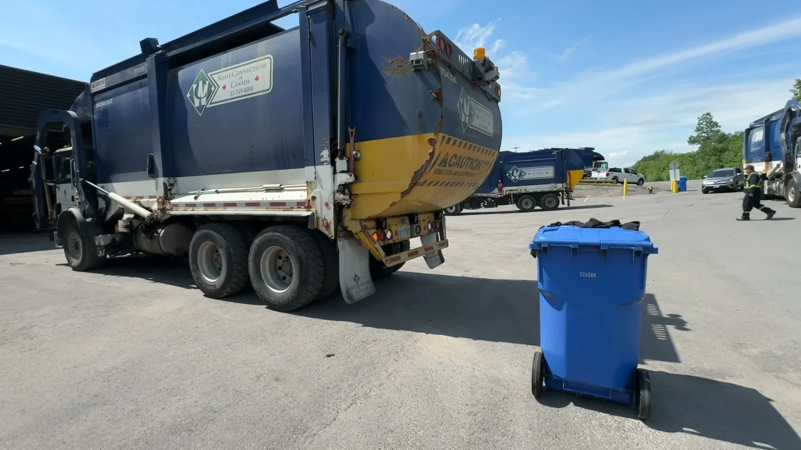 Waste Connections front-load truck reversing toward a blue recycling cart in a yard